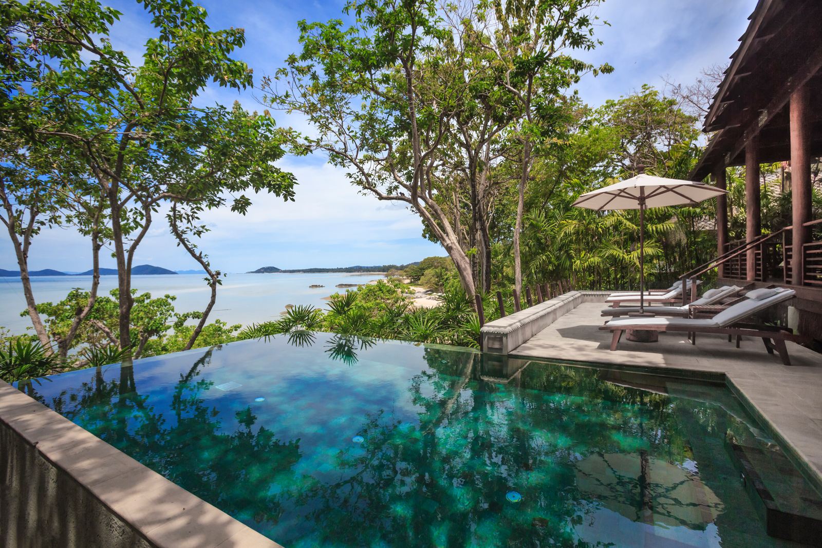 An infinity pool overlooking a serene tropical coastline, surrounded by lush trees and greenery with a stunning ocean and distant islands in the background. The pool area features comfortable loungers and an elegant open-air design.