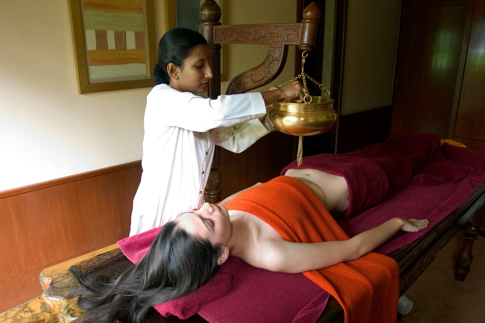 A practitioner performs a traditional Ayurvedic oil treatment using a suspended brass bowl over a relaxed client on a treatment table. The scene depicts a calm, therapeutic wellness environment.