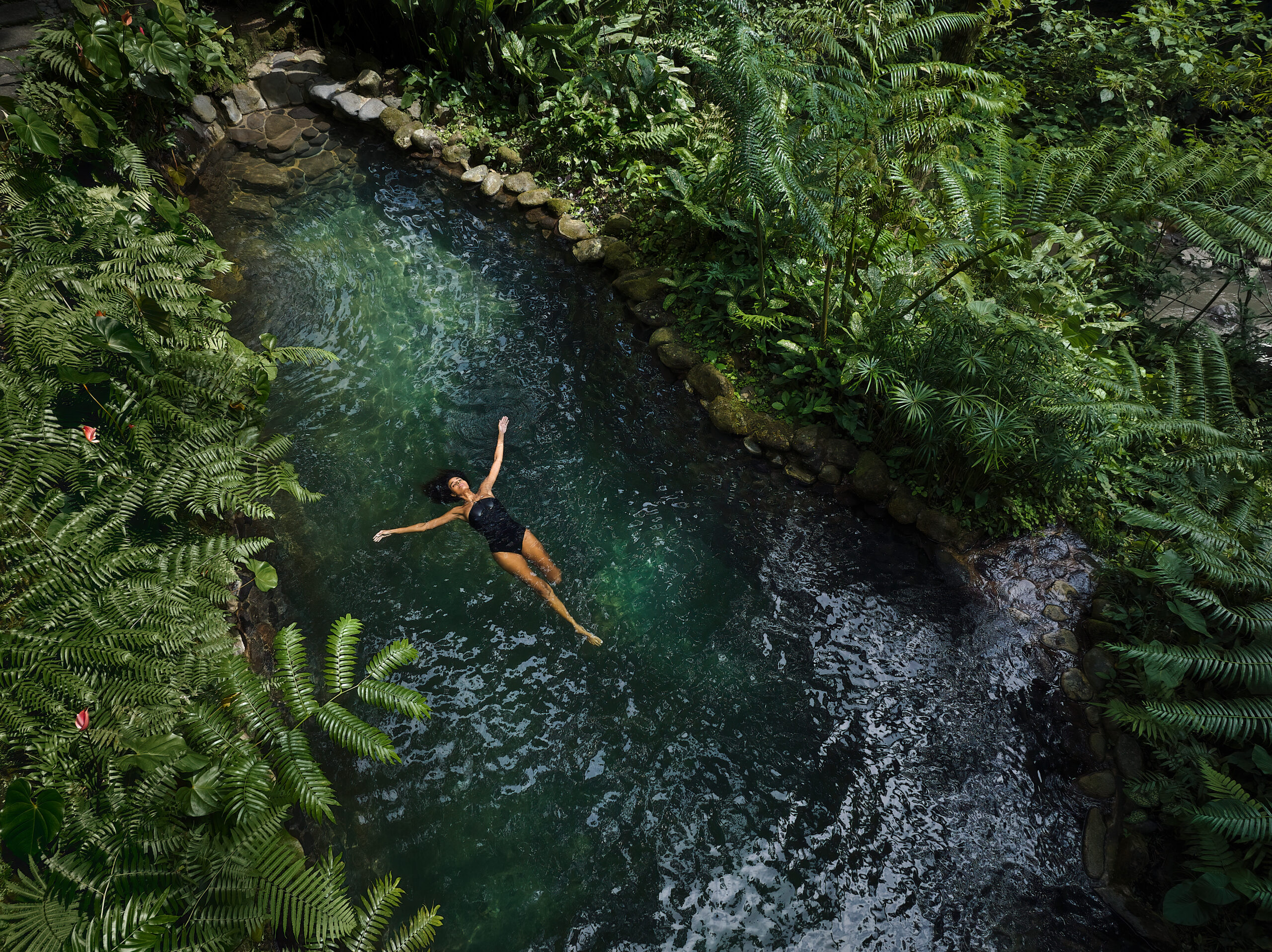 A person floating peacefully in a lush green jungle stream surrounded by ferns and tropical vegetation. The water is a deep emerald green with stone-lined banks.