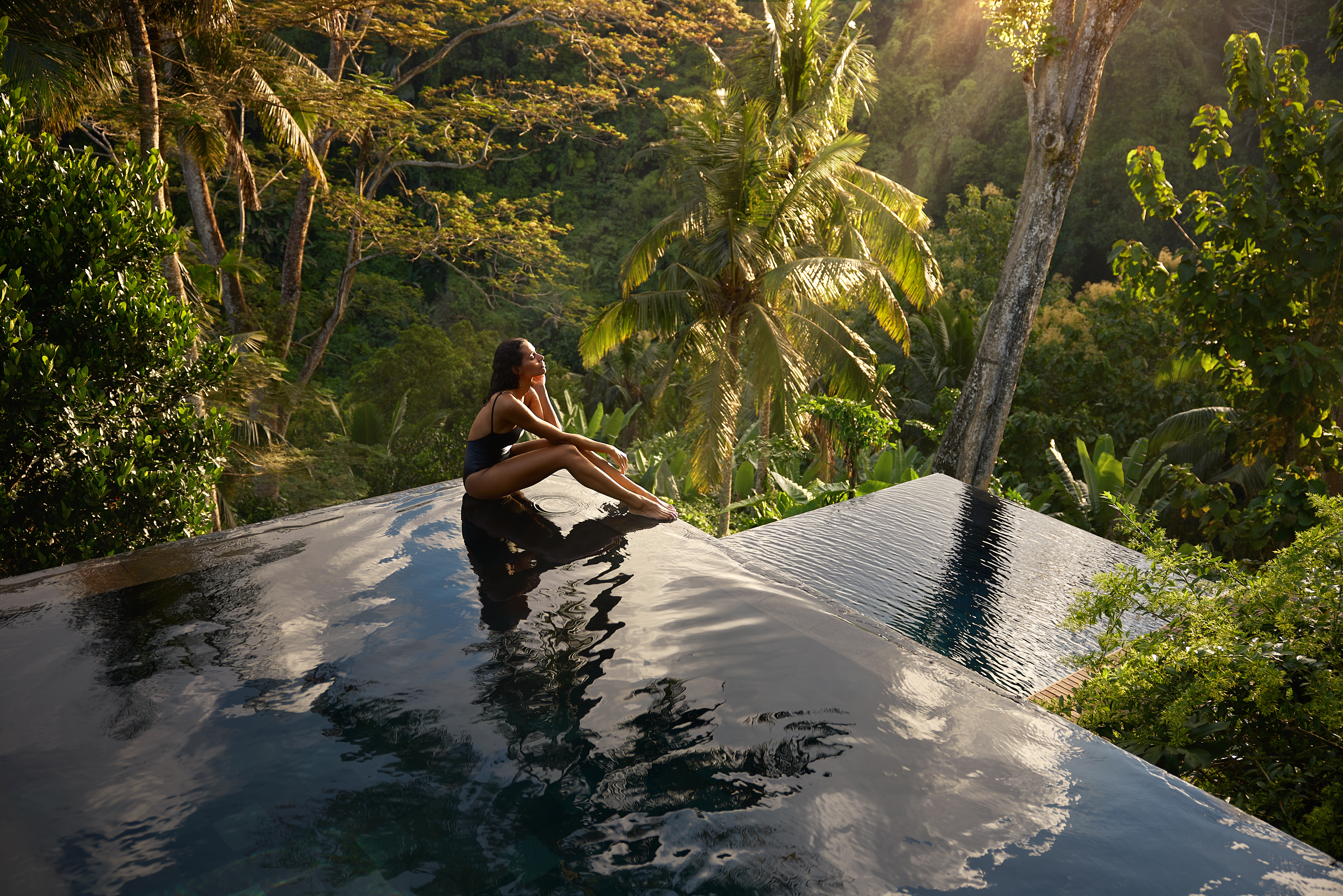 A serene infinity pool overlooking a dense tropical forest with palm trees and sunlight filtering through the foliage. A person sits peacefully at the edge of the pool, enjoying the tranquil landscape.