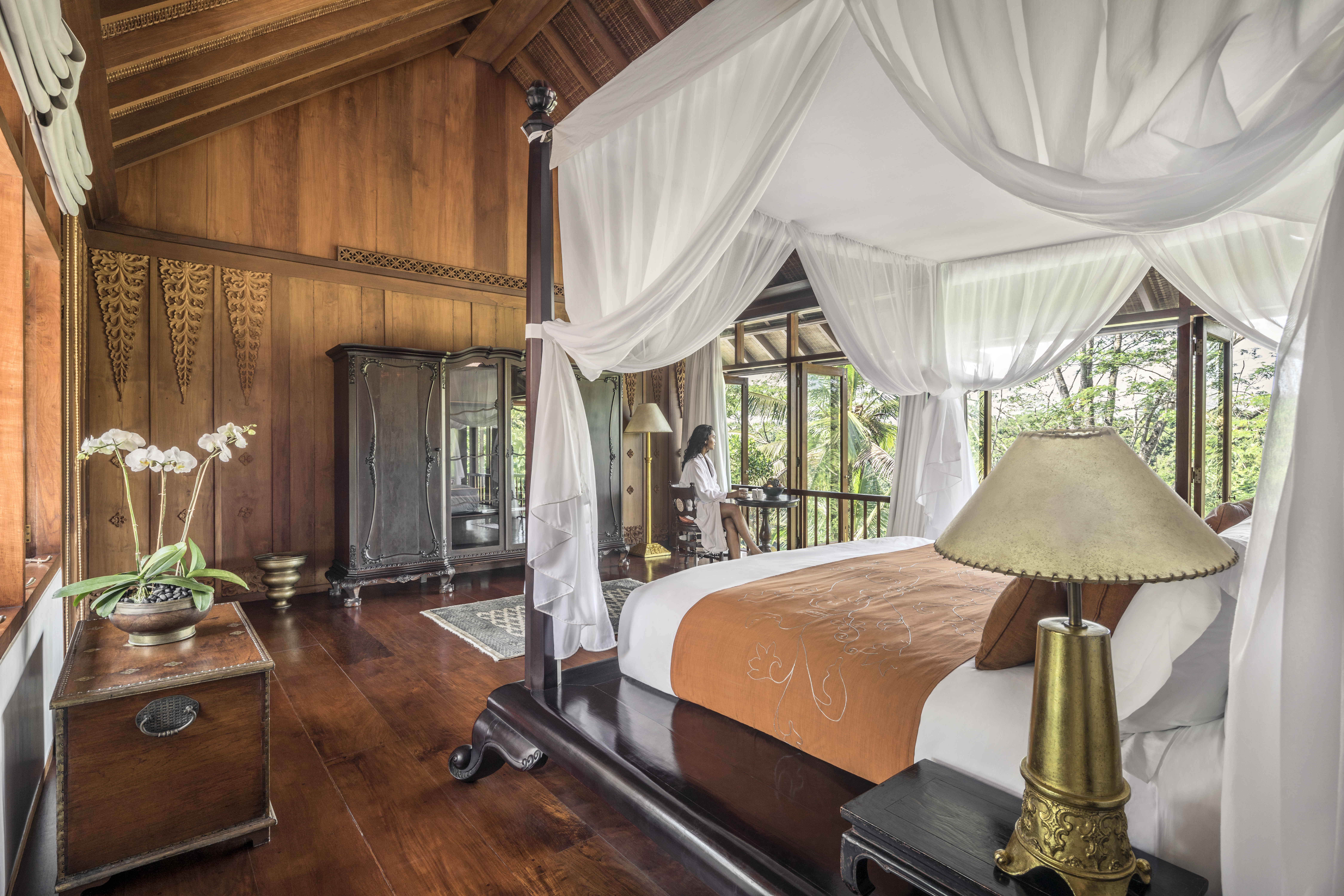 An elegant wooden-paneled bedroom with a canopy bed, featuring white curtains and a view of lush tropical surroundings. The room combines traditional Balinese architectural elements with modern luxury resort design.