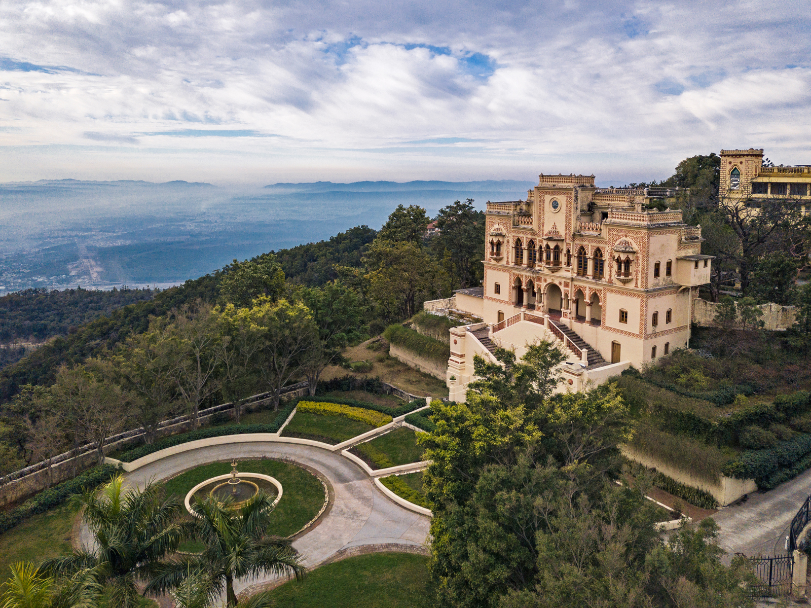 An elegant, ornate European-style castle situated on a forested hilltop with sweeping views of a misty valley and distant mountain ranges. The castle features intricate architectural details and is surrounded by manicured gardens with a central circular fountain.