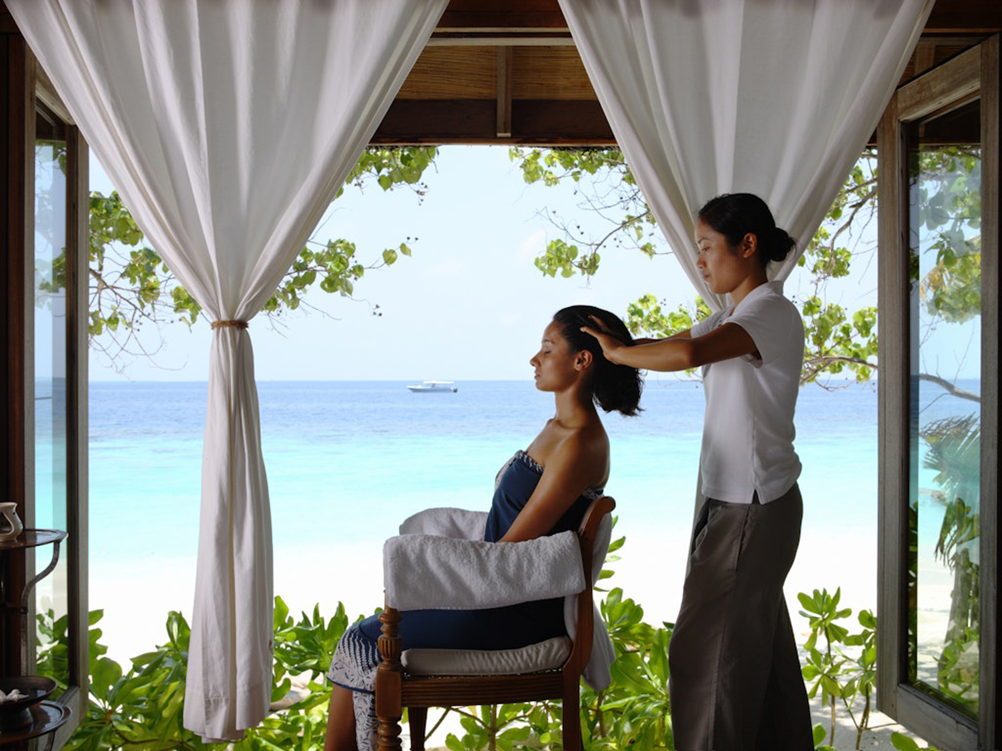 Indian_Head_Massage_3_Medium A woman in a white bathrobe sits on a cushioned bench near elegant sculptural lighting, creating a calm and luxurious wellness environment.