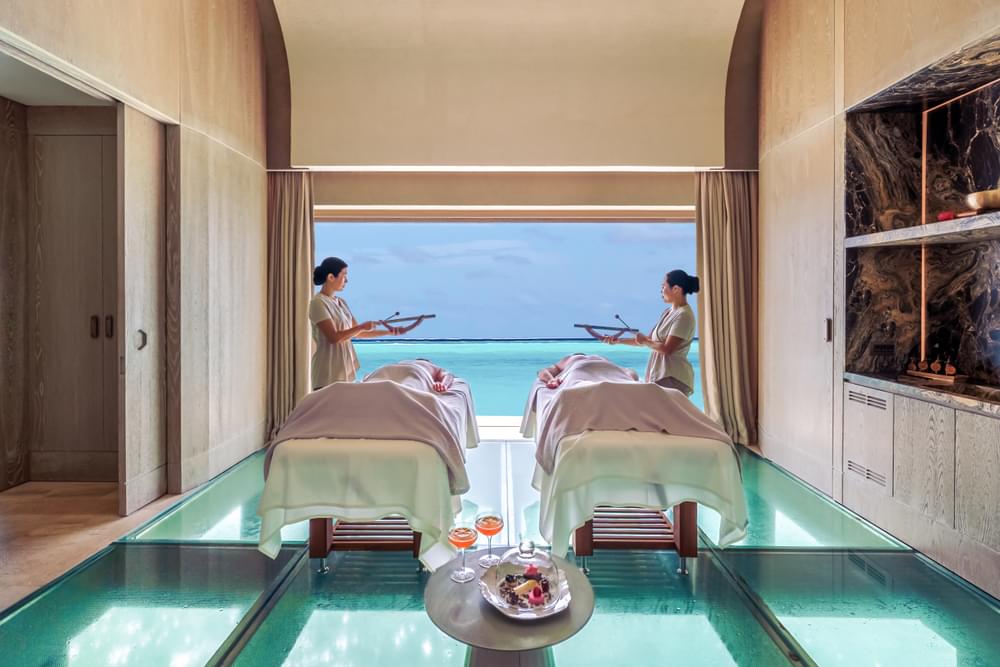 A circular logo sign for the Joali Being sea turtle rehabilitation center, featuring a stylized blue wave design and located in a tropical outdoor setting with plants and wooden fencing