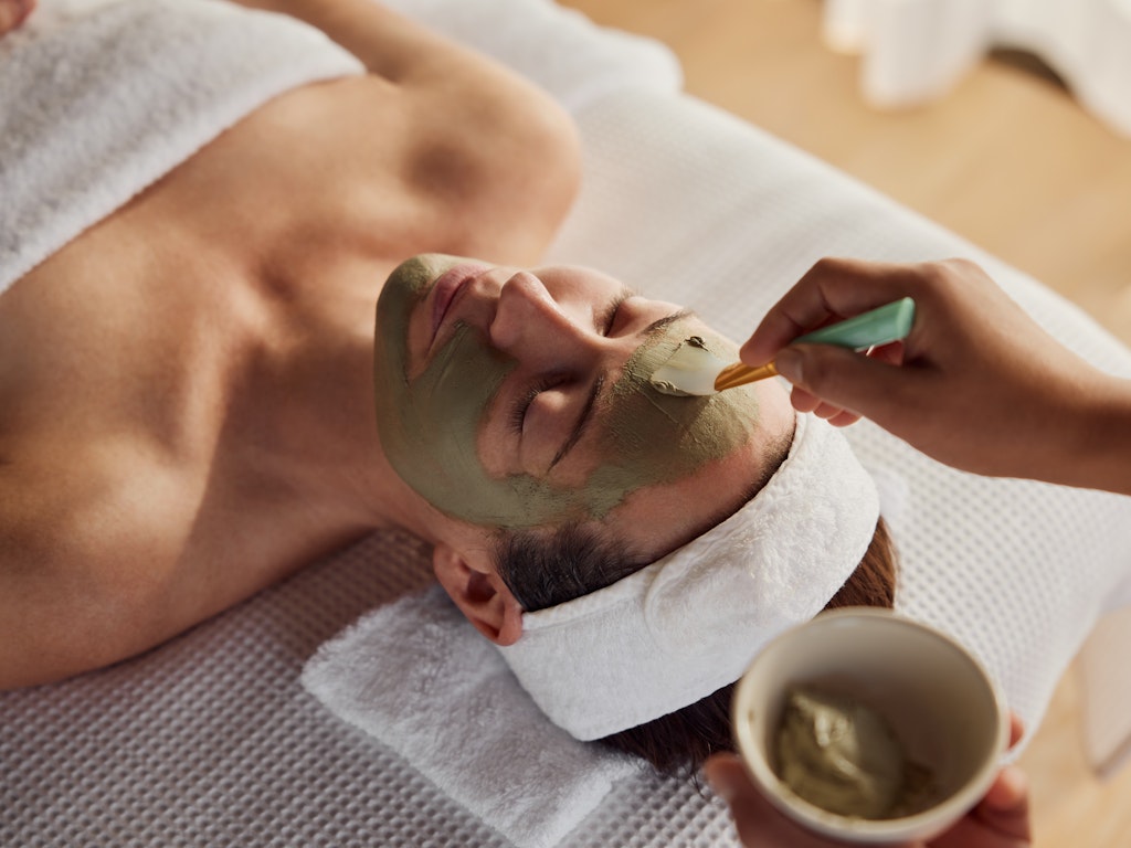 A luxurious traditional Balinese-style bedroom with a four-poster bed draped in white mosquito netting, featuring a thatched roof and stone wall elements.
