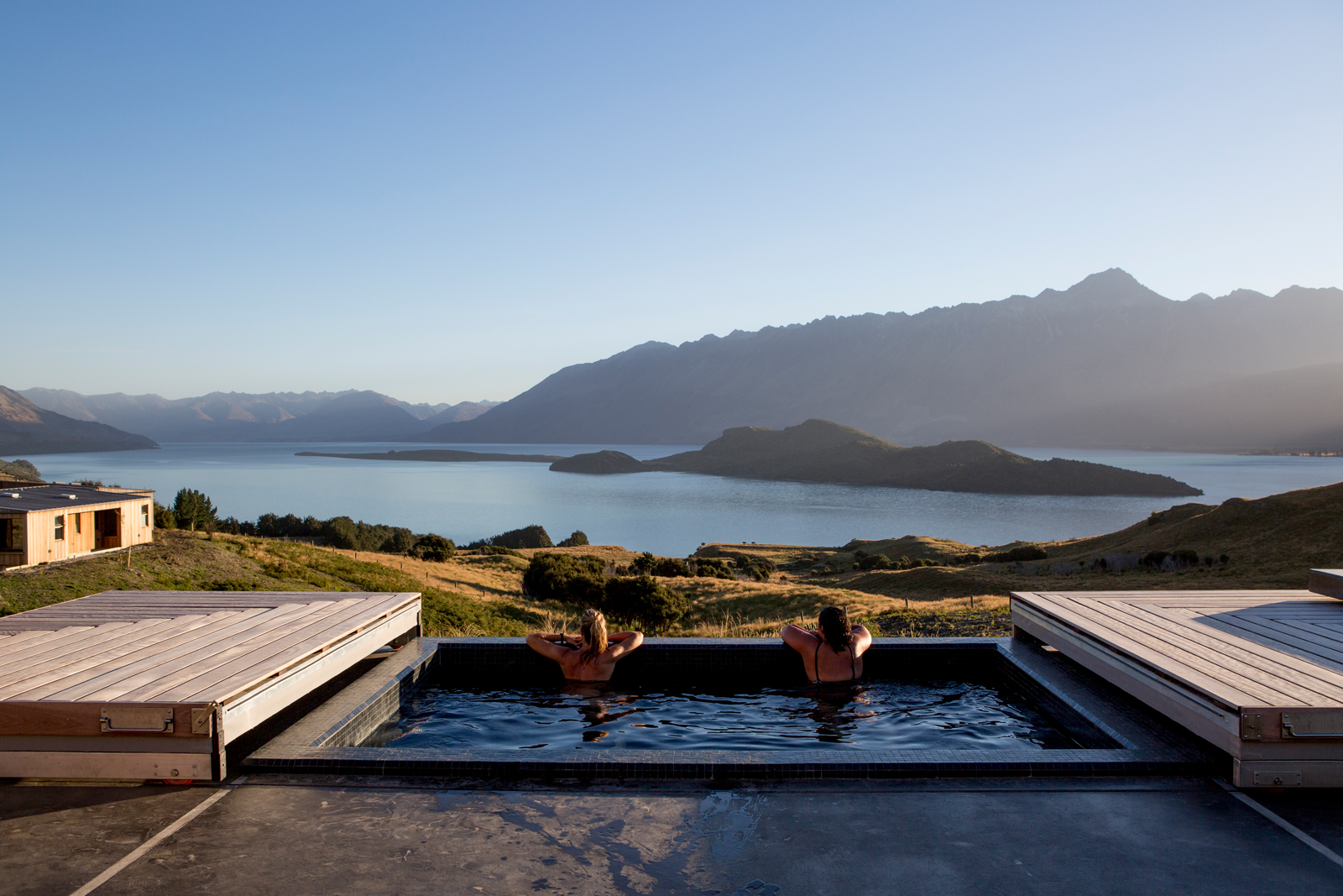Two people practicing yoga on pink mats near a swimming pool with hanging ferns and wicker furniture. The setting appears to be a private, elegant outdoor space with stone flooring and a water feature.