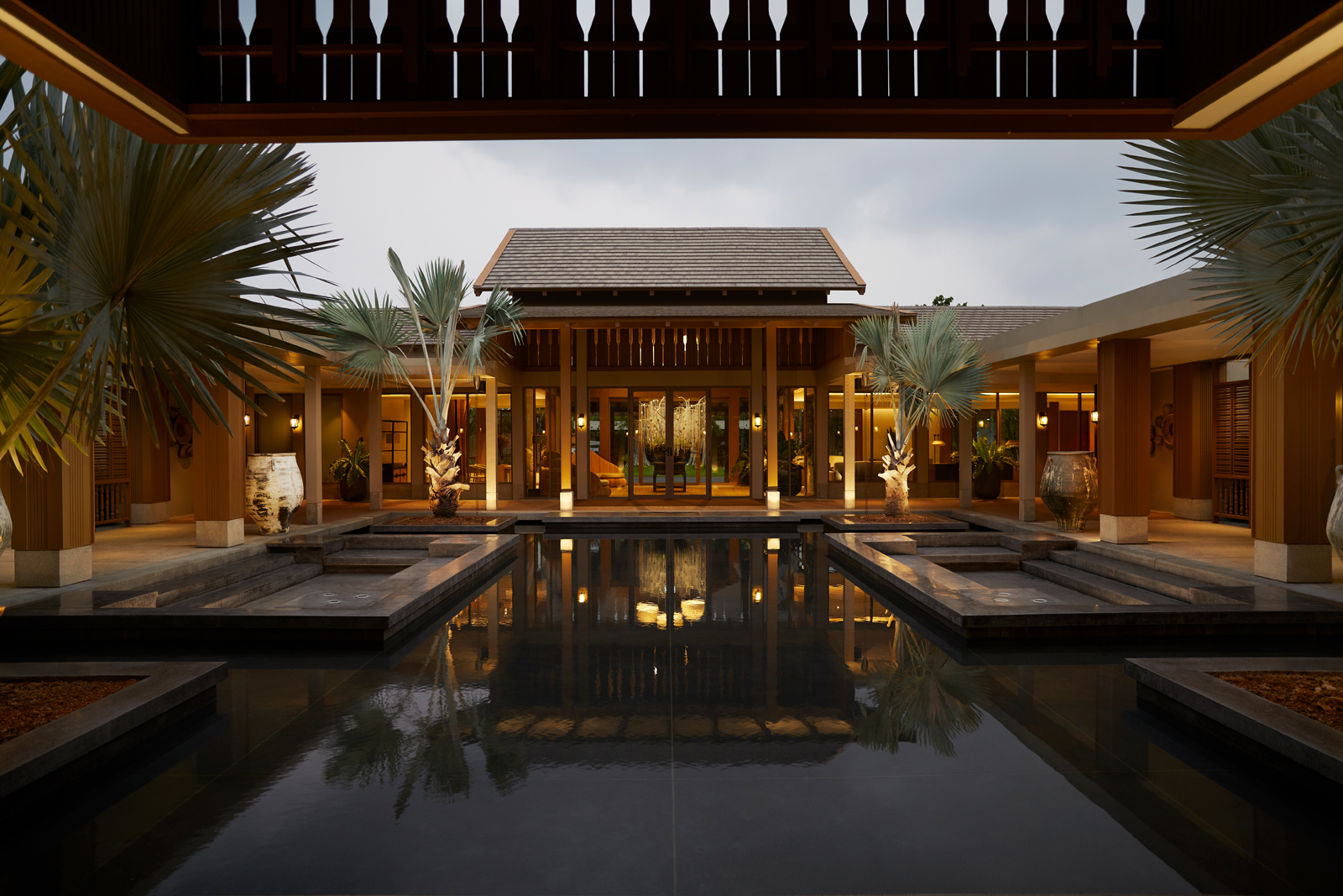 An elegant tropical resort architectural design featuring a central pavilion with symmetrical wings, surrounded by palm trees and a serene reflection pool illuminated during twilight hours.