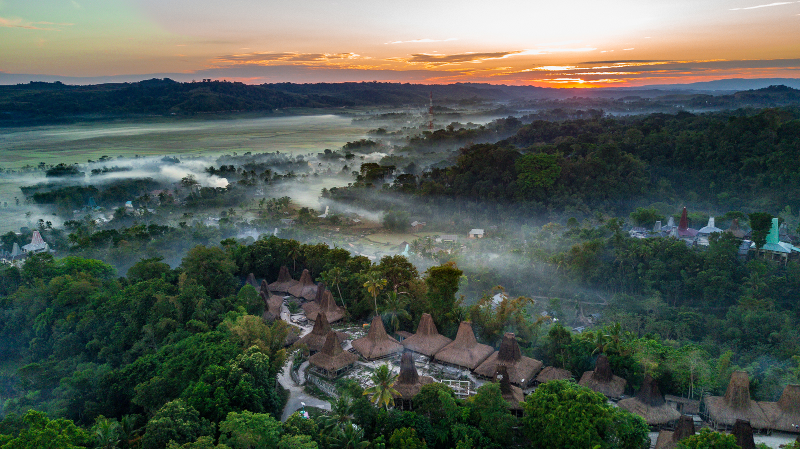 A breathtaking aerial view of a traditional village with thatched-roof houses nestled in a lush, misty tropical landscape at sunrise, showcasing the beautiful natural scenery and cultural architecture of Indonesia.
