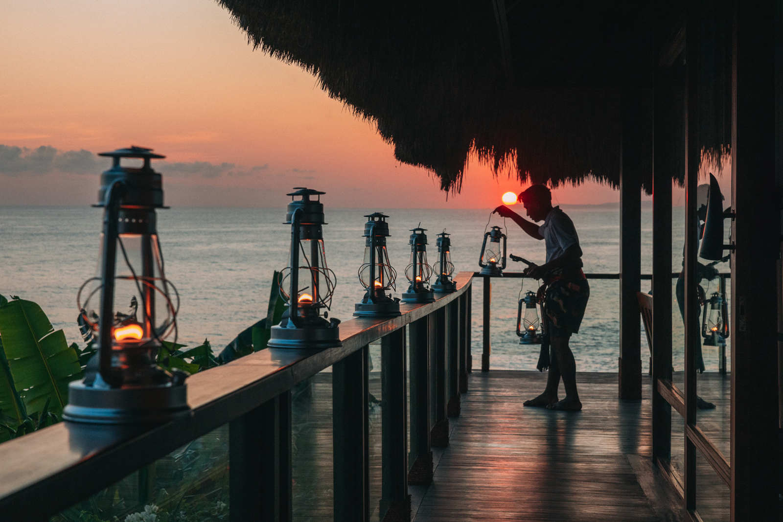 A serene scene of vintage lanterns being lit on a wooden pier at sunset, with a thatched roof overhead and the ocean stretching to the horizon.