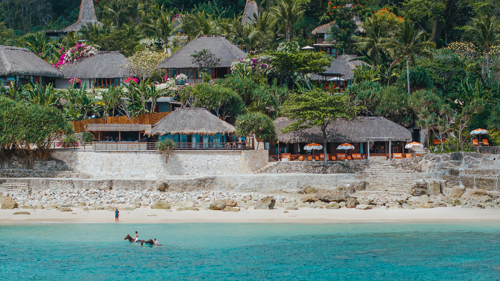 A luxurious seaside resort with traditional thatched-roof bungalows nestled among lush tropical greenery, overlooking a pristine turquoise bay with rocky shoreline and white sandy beach.