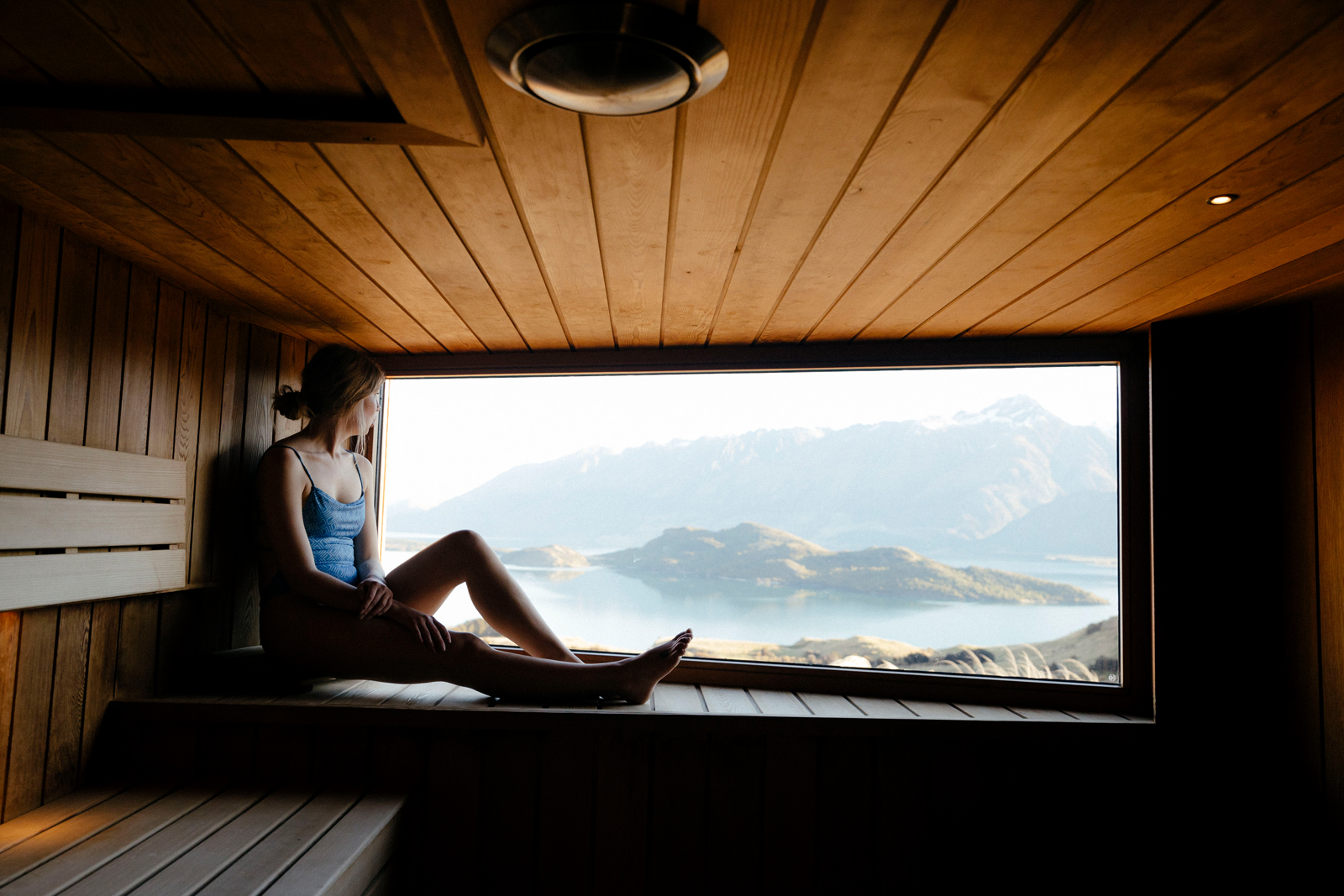 A person relaxes in a wooden sauna with a large panoramic window overlooking a scenic mountain landscape with a lake and misty terrain.