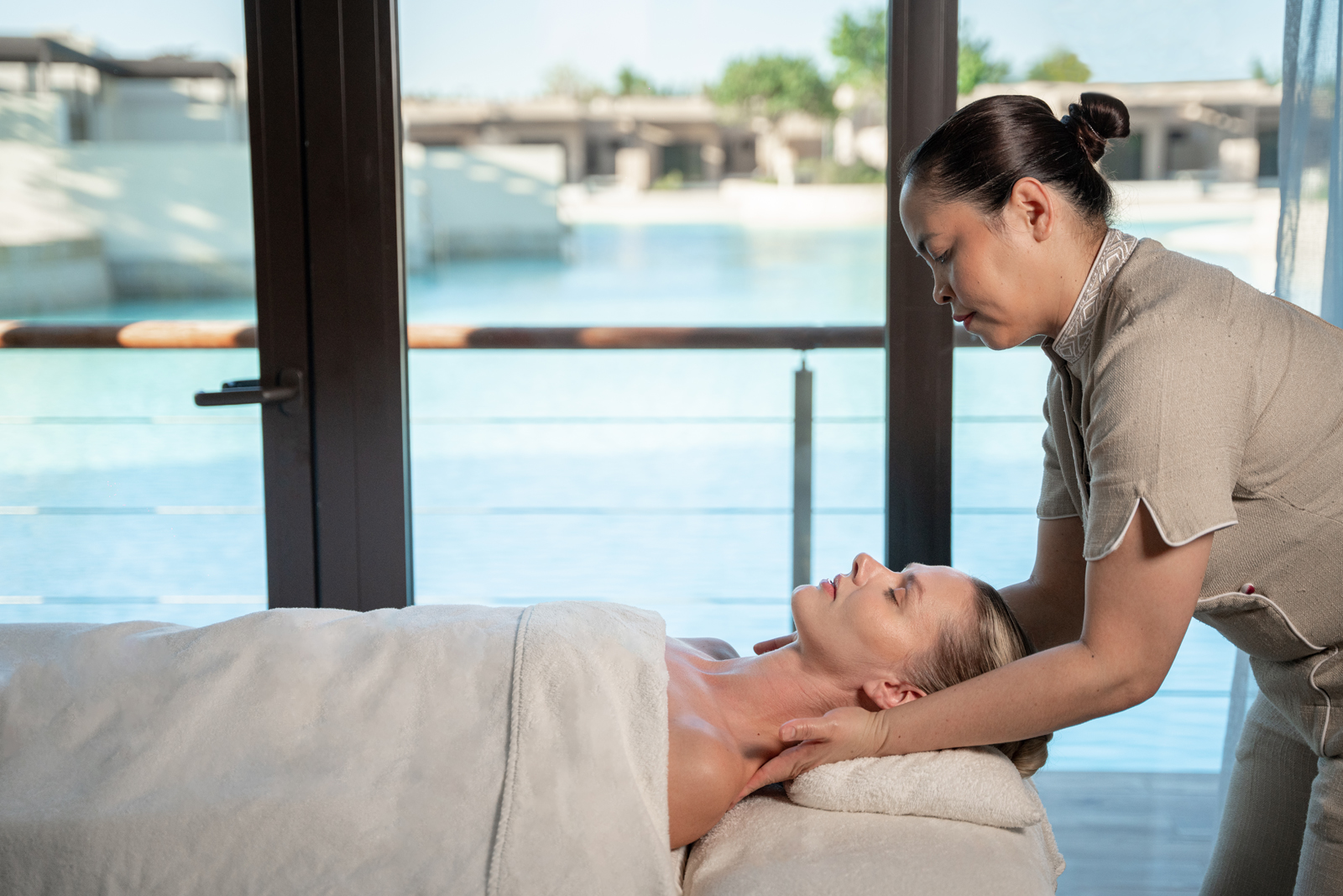A professional spa therapist performing a massage treatment for a client in a serene wellness space with a swimming pool visible through large windows.