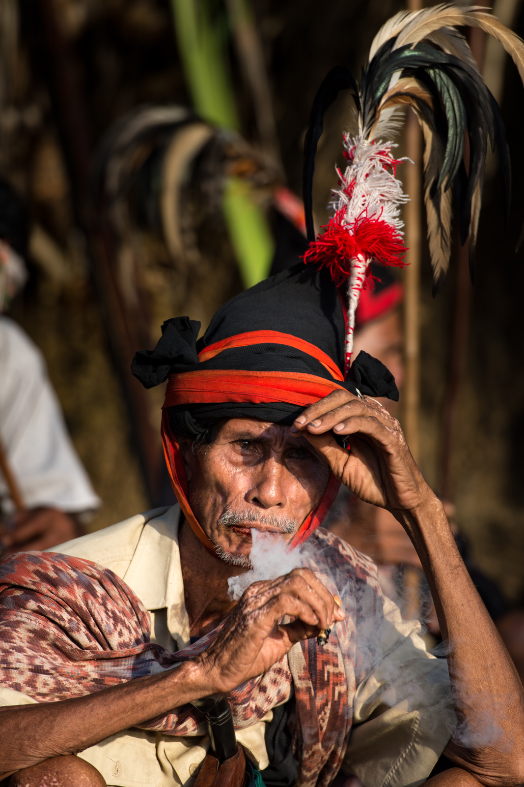 An elderly indigenous performer wearing a distinctive black and red headdress with feathers, smoking and looking contemplative during a cultural event or ceremony.