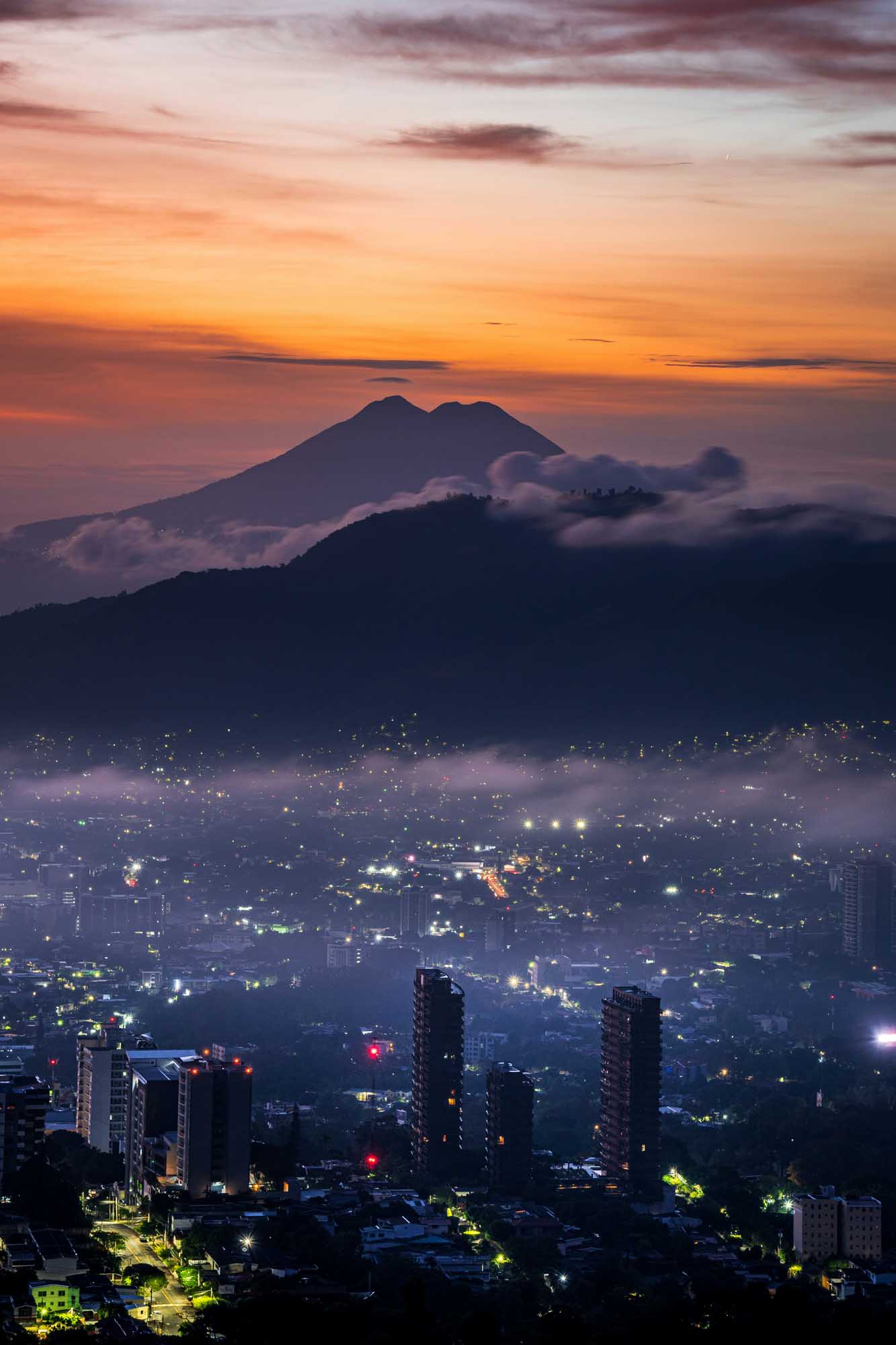 A stunning twilight cityscape with a dramatic volcanic mountain silhouetted against an orange